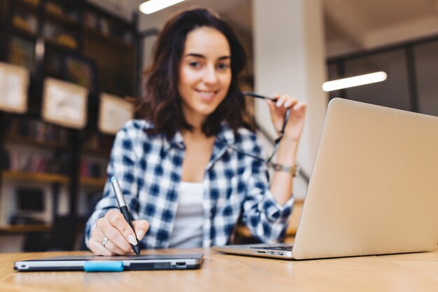 Immagine moderna ed elegante della giovane donna castana intelligente che lavora con il computer portatile sul tavolo in libreria. Sorridente, gioco con occhiali neri, grande successo, studente laborioso.