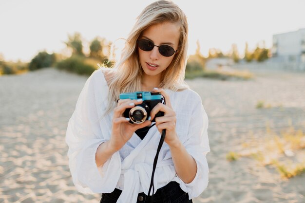 Immagine positiva di stile di vita della ragazza bionda alla moda divertendosi e facendo foto sulla spiaggia vuota. Vacanze e tempo di ferie. Libertà e natura in campagna.