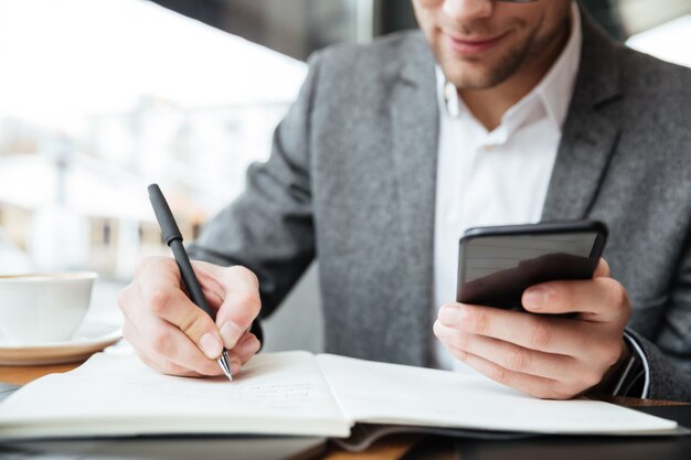 Immagine potata dell'uomo d'affari calmo che si siede dalla tavola in caffè mentre usando smartphone e scrivendo qualcosa