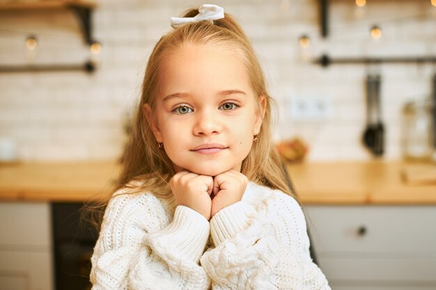 Immagine ravvicinata di adorabile bambina con gli occhi verdi e lunghi capelli sciolti che tengono le mani sotto il mento con il sorriso in posa in cucina