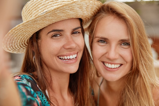 Immagine ravvicinata di adorabili femmine hanno relazioni amichevoli, fanno selfie, hanno ampi sorrisi, stanno vicine l'una all'altra. Le coppie omosessuali ricreano insieme sul resort tropicale. Concetto di positività