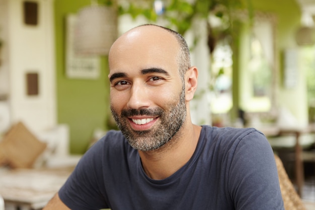 Immagine ravvicinata di bell'uomo caucasico con la barba vestito in t-shirt alla ricerca e sorridente con felice espressione allegra, seduto al ristorante sul marciapiede in giornata di sole, in attesa di amici