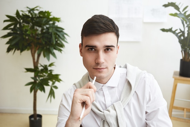 Immagine ravvicinata di bello senza problemi giovane brunetta uomo dipendente che indossa abiti formali eleganti con sguardo pensieroso, tenendo la matita, seduto in ufficio interno