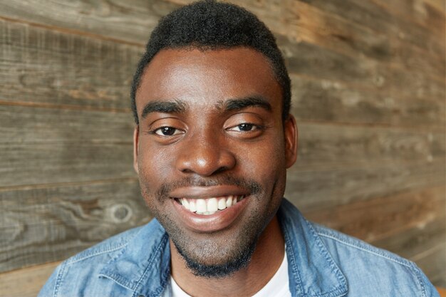 Immagine ravvicinata di bello studente africano con la barba vestita in camicia di jeans che sorride felicemente, mostrando i suoi denti bianchi, con un aspetto gioioso e contento