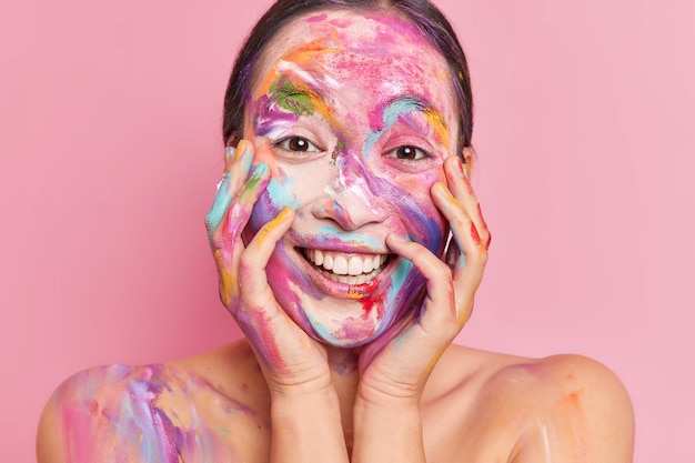 Immagine ravvicinata di donna asiatica positiva spalmata di vernici colorate a guazzo tiene le mani sulle guance sorrisi felicemente