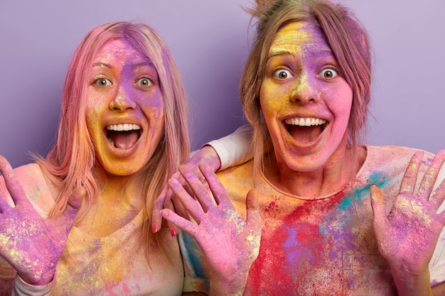 Immagine ravvicinata di due donne emotive felicissime che aprono la bocca, esclamano con gioia, hanno colorato la festa, si sono spalmate a vicenda con polvere colorata, stupite nel vedere qualcosa di incredibile sul festival indiano
