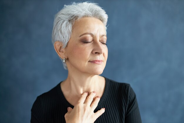 Immagine ravvicinata di elegante attraente donna di mezza età con capelli grigi e rughe, chiudendo gli occhi e sorridendo, toccando il collo, applicando crema antietà