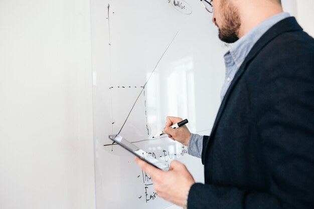 Immagine ravvicinata di giovane uomo dai capelli scuri in bicchieri scrivendo un business plan sulla lavagna Vista laterale, messa a fuoco a portata di mano.