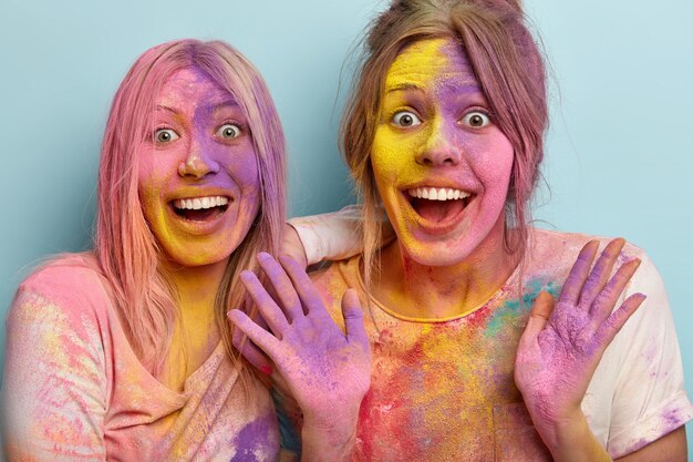 Immagine ravvicinata di giovani donne felici che si divertono con polvere colorata al festival di Holi, sorridono ampiamente, hanno facce multicolori, ascoltano incredibili buone notizie, isolate su un muro blu. Giorno festivo