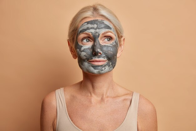 Immagine ravvicinata di premurosa donna europea applica maschera facciale di argilla e distoglie lo sguardo si preoccupa per la carnagione pone con le spalle nude isolate sulla parete marrone