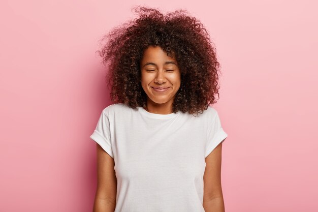 Immagine ravvicinata di un adorabile adolescente soddisfatto con i capelli ricci e folti, ha gli occhi chiusi, un sorriso piacevole, attende la sorpresa con grande felicità, si gode un momento fantastico durante il fine settimana, indossa una maglietta bianca