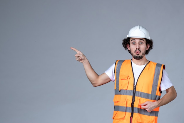 Immagine ravvicinata di un architetto maschio emotivo scioccato in giubbotto di avvertimento con casco di sicurezza e che punta qualcosa sopra la parete grigia dell'onda