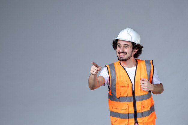 Immagine ravvicinata di un architetto maschio sorridente in giubbotto di avvertimento con casco di sicurezza e puntato in avanti sopra la parete grigia dell'onda