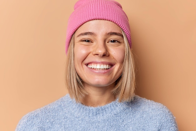 Immagine ravvicinata di una giovane donna dai capelli biondi felice che mostra ampiamente i denti bianchi perfetti che sono di buon umore indossa un maglione casual e un cappello isolati su sfondo marrone. Concetto di emozioni positive