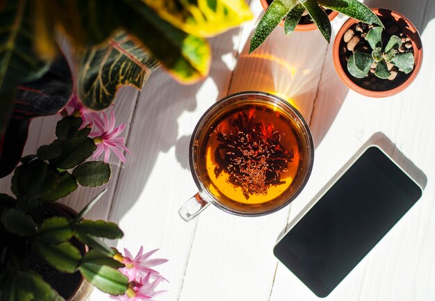 Immagine ravvicinata di una tazza di tè in vetro, fragole rosse, smartphone e fiori in vasi su uno sfondo di legno chiaro.