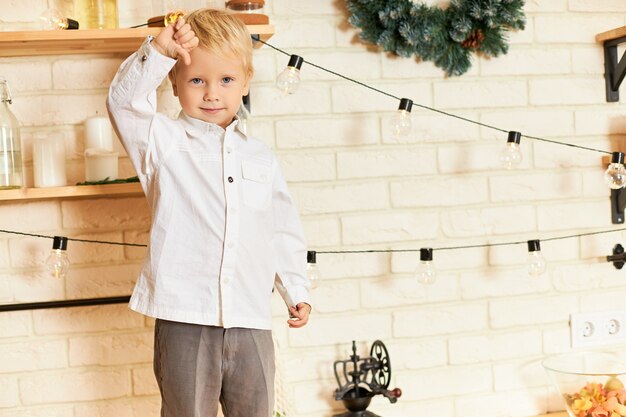 Immagine ritagliata del bambino maschio europeo biondo carino che indossa una camicia bianca in posa a piedi nudi in cucina, in piedi sul bancone, esprimendo antipatia o reazione negativa, mostrando il gesto del pollice verso il basso