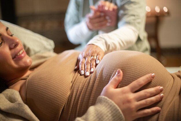 Immagine ritagliata di donna incinta sorridente e ostetrica a casa. Donna in abiti casual sdraiato sul letto, ostetrica che tiene la mano. Gravidanza, medicina, concetto di nascita in casa