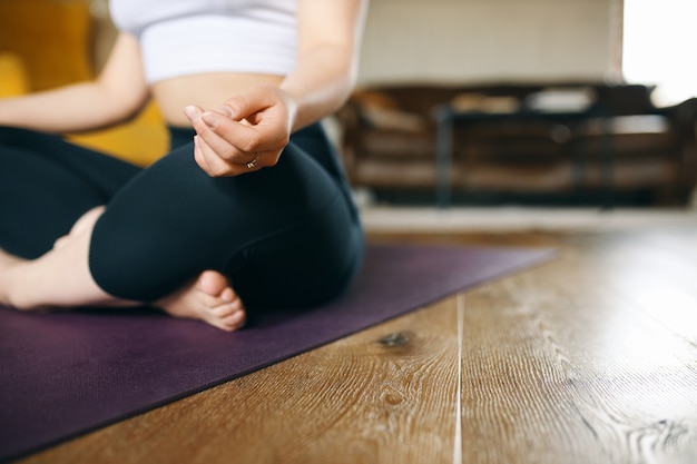 Immagine ritagliata di giovane femmina muscolosa in forma in abiti sportivi che medita sul pavimento nella posa di mezzo loto, facendo il gesto del mudra, seduto sulla stuoia prima della pratica dello yoga, concentrandosi sui sentimenti e sulla respirazione