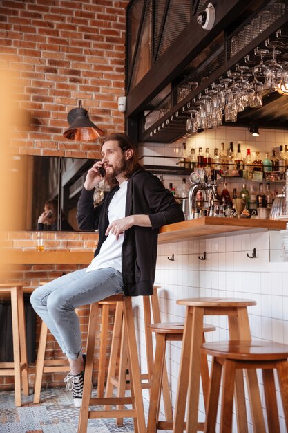 Immagine verticale dell'uomo barbuto che si siede sulla barra con il telefono