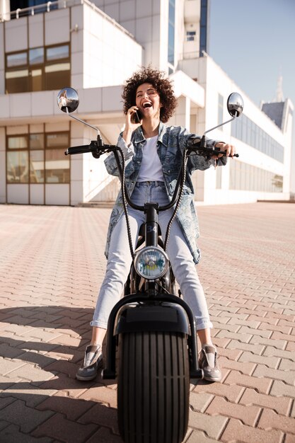 Immagine verticale della donna riccia felice che si siede sulla motocicletta moderna