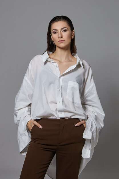 Immagine verticale della giovane donna europea fiduciosa alla moda con i capelli scuri pettinati all'indietro in posa, indossa pantaloni marroni eleganti e camicia bianca oversize, tenendo le mani in tasca