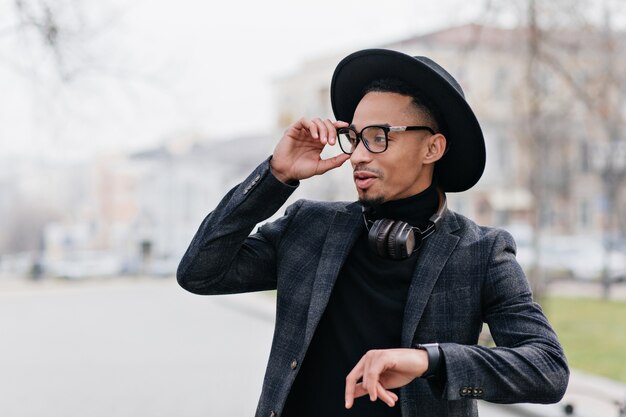 Impegnato uomo africano in elegante cappello guardandosi intorno attraverso gli occhiali. Ritratto di modello maschio romantico in abito nero in posa sulla città di sfocatura.