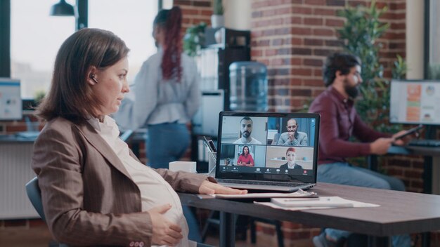 Impiegata incinta che utilizza la videochiamata aziendale sul laptop per parlare con i colleghi. Donna che aspetta un bambino e partecipa alla riunione in videoconferenza remota online, parlando della pianificazione del progetto.