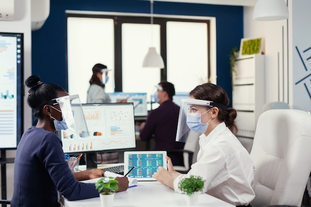 Impiegato africano che discute con il manager seduto alla scrivania sul posto di lavoro che indossa una maschera facciale contro il covid19. Diversi gruppi di uomini d'affari che lavorano e comunicano insieme in un ufficio creativo con nuovi