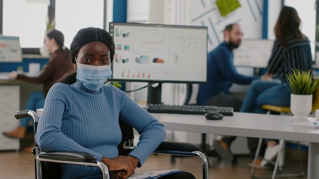 Impiegato africano disabile paralizzato immobilizzato che guarda la telecamera indossando una maschera di protezione durante ...