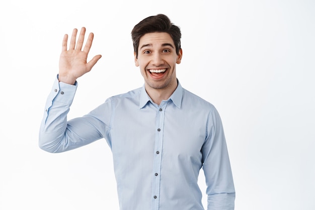 Impiegato amichevole agitando la mano e salutando, sorridendo saluta, salutando, dando un caloroso benvenuto e guardando allegro, in piedi contro il muro bianco