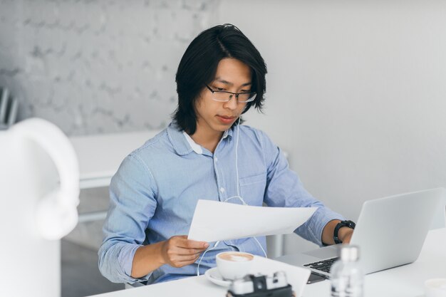Impiegato asiatico concentrato in auricolari che leggono documenti sul posto di lavoro. Il ritratto dell'interno dello specialista it freelance cinese beve il caffè mentre utilizza con il computer portatile.