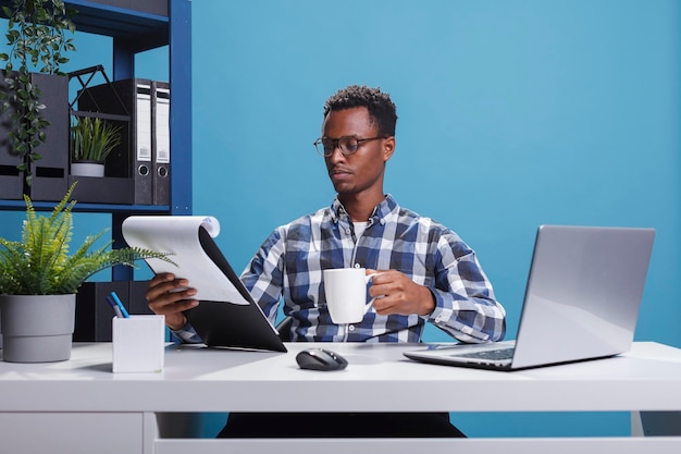 Impiegato aziendale con appunti di documentazione e tazza di caffè che esaminano i grafici di gestione del progetto. Giovane impiegato afroamericano seduto in uno spazio di lavoro moderno mentre esamina le scartoffie.