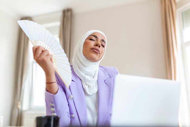 Impiegato dell'ufficio arabo che soffre di aria calda e soffocante che lavora al computer portatile da casa senza condizionatore nei giorni estivi Donna d'affari musulmana che si raffredda con un ventilatore portatile sul posto di lavoro