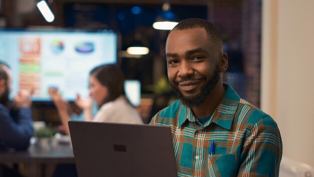 Impiegato di ufficio afroamericano sorridente che lavora al ritratto del computer portatile. Giovane che tiene il computer, guardando la telecamera da vicino, presentazione finanziaria dell'azienda, sfondo della riunione di lavoro. Tiro a mano libera.
