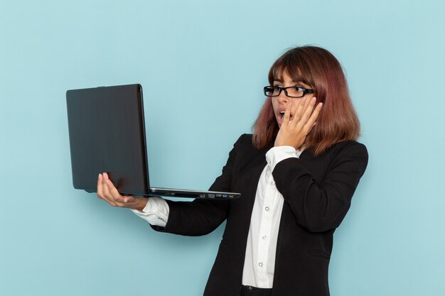 Impiegato di ufficio femminile di vista frontale in vestito rigoroso utilizzando il computer portatile con l'espressione scioccata sulla superficie azzurra