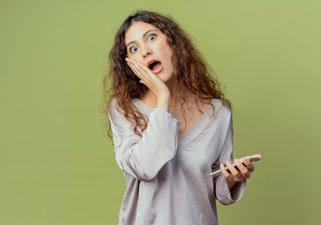 Impiegato di ufficio femminile giovane sorpreso che tiene il telefono e che mette la mano sulla guancia isolata sulla parete verde oliva