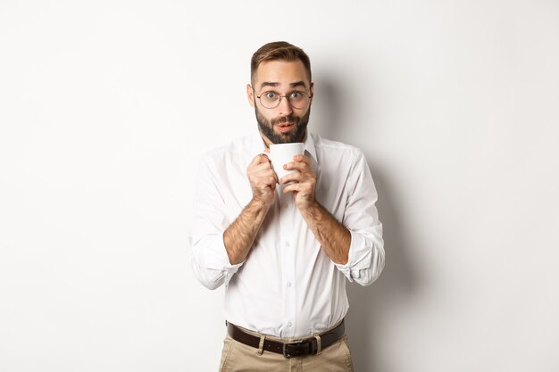 Impiegato felice che beve caffè caldo e sembra eccitato, spettegolare, in piedi