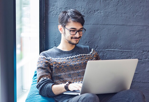 Impiegato maschio che si siede su un sofà accanto ad una finestra con il suo computer portatile