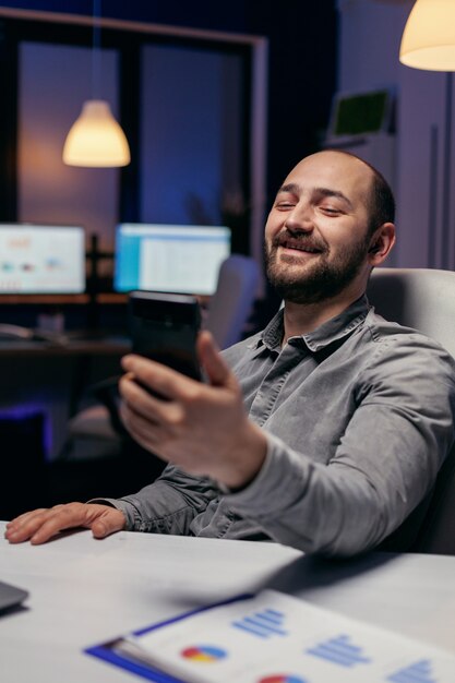 Imprenditore allegro che saluta parlando in videochiamata usando il telefono. Imprenditore nel corso di un'importante videoconferenza mentre fa gli straordinari in ufficio.