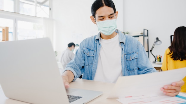 Imprenditore asiatico uomo d'affari che indossa una maschera medica per l'allontanamento sociale in una nuova situazione normale per la prevenzione dei virus mentre si utilizza il laptop al lavoro in ufficio. Stile di vita dopo il virus corona.