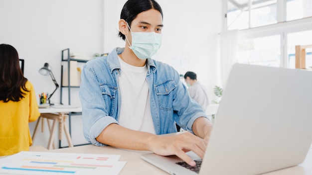 Imprenditore asiatico uomo d'affari che indossa una maschera medica per l'allontanamento sociale in una nuova situazione normale per la prevenzione dei virus mentre si utilizza il laptop al lavoro in ufficio. Stile di vita dopo il virus corona.