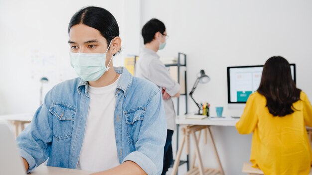 Imprenditore asiatico uomo d'affari che indossa una maschera medica per l'allontanamento sociale in una nuova situazione normale per la prevenzione dei virus mentre si utilizza il laptop al lavoro in ufficio. Stile di vita dopo il virus corona.