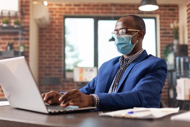 Imprenditore che guarda lo schermo del laptop per lavorare su un progetto aziendale durante la pandemia. Dipendente che indossa una maschera facciale e lavora al computer per pianificare la strategia di marketing nell'ufficio di avvio.