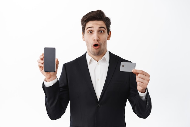 Imprenditore impressionato che mostra telefono e carta di credito ansimante stupito in piedi su sfondo bianco in abito nero