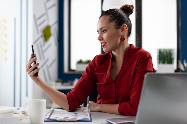 Imprenditore in videoconferenza casuale tramite smartphone. Donna di affari sorridente in camicia rossa che utilizza il telefono cellulare nell'ufficio di affari di avvio. Startup manager che esamina i contenuti sui social media.
