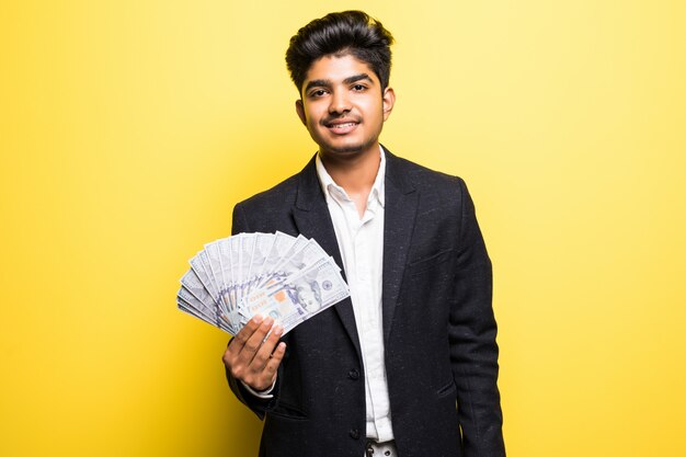 Imprenditore indiano di successo con il vestito classico disponibile delle banconote del dollaro che esamina macchina fotografica con il sorriso a trentadue denti mentre stando contro la parete gialla