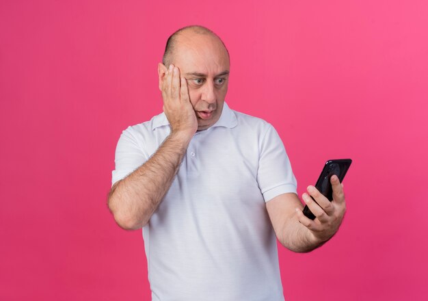 Imprenditore maturo casual sorpreso tenendo e guardando il telefono cellulare e tenendo la mano sul viso isolato su sfondo rosa con spazio di copia