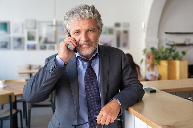 Imprenditore maturo di successo parlando al telefono cellulare, in piedi al co-working, appoggiato sulla scrivania, guardando la fotocamera a