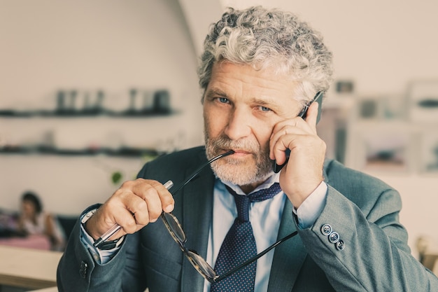 Imprenditore maturo pensieroso parlando al telefono cellulare, in piedi al co-working, appoggiato sulla scrivania