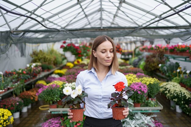 Imprenditore sorridente nel suo vivaio in piedi tenendo in mano due vasi con fiori rossi e bianchi nella serra